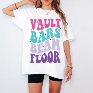 May include: White oversized t-shirt with the text "VAULT BARS BEAM FLOOR" in a retro font with pink, blue, and purple colors.