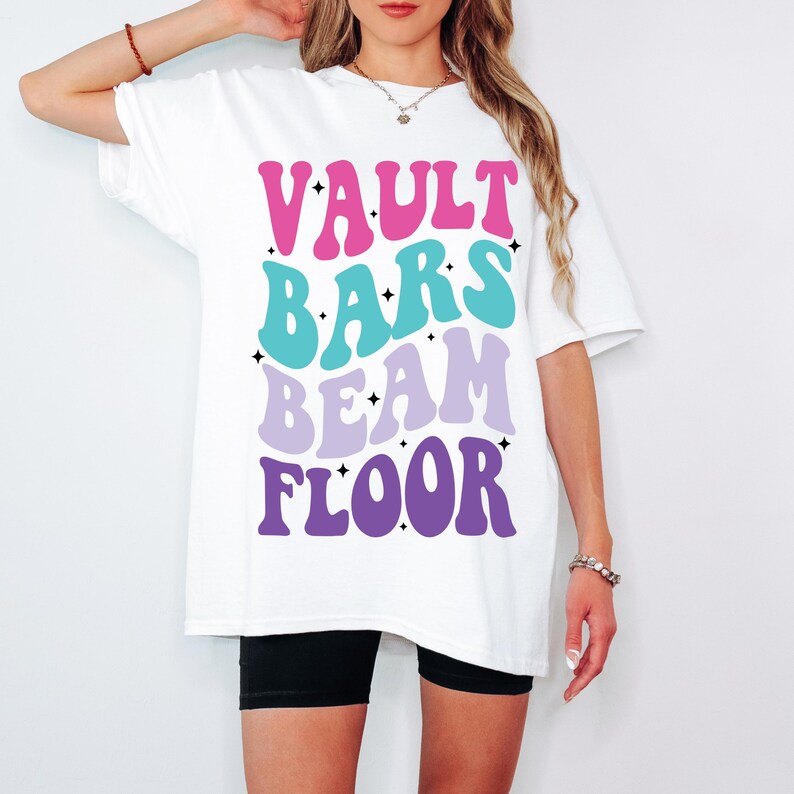 May include: White oversized t-shirt with the text "VAULT BARS BEAM FLOOR" in a retro font with pink, blue, and purple colors.