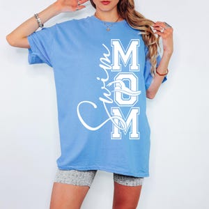 May include: A light blue t-shirt with the text "Swim Mom" in white lettering. The "Mom" is in a varsity style font with a swimmer graphic in the "O".