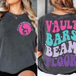 May include: A dark gray t-shirt with a pink circle and a silhouette of a gymnast doing a handstand. The back of the shirt has the words "VAULT BARS BEAM FLOOR" in a retro font with pink, blue, and purple colors.