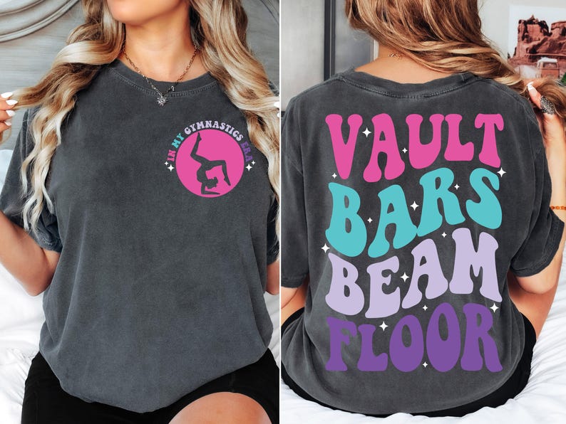 May include: A dark gray t-shirt with a pink circle and a silhouette of a gymnast doing a handstand. The back of the shirt has the words "VAULT BARS BEAM FLOOR" in a retro font with pink, blue, and purple colors.