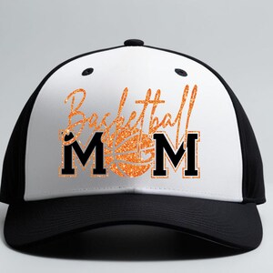 May include: A black and white baseball cap with the words "Basketball Mom" in orange glitter script. The word "MOM" is in bold black letters. A basketball graphic is in the center of the text.