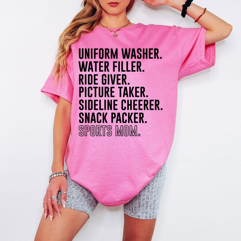 May include: Pink t-shirt with black text that reads "UNIFORM WASHER. WATER FILLER. RIDE GIVER. PICTURE TAKER. SIDELINE CHEERER. SNACK PACKER. SPORTS MOM."