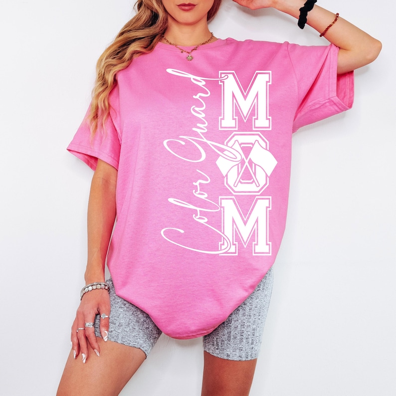 May include: Pink t-shirt with white text that reads "Color Guard MOM" with a graphic of crossed flags.