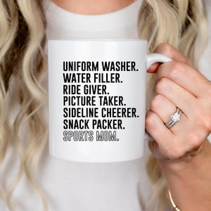 May include: White ceramic mug with black text that reads "Uniform Washer. Water Filler. Ride Giver. Picture Taker. Sideline Cheerer. Snack Packer. Sports Mom."