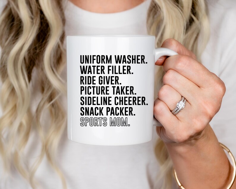 May include: White ceramic mug with black text that reads "Uniform Washer. Water Filler. Ride Giver. Picture Taker. Sideline Cheerer. Snack Packer. Sports Mom."