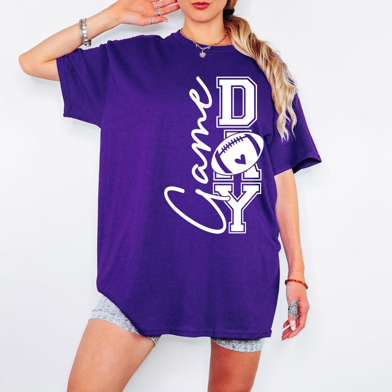 May include: A purple t-shirt with the words "Game Day" in a white script and block letters. A white football with a heart is in the center of the "Day" text. The shirt is a short-sleeved, oversized style.