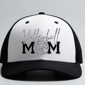 May include: A black and white baseball cap with the words "Volleyball Mom" in silver glitter script and block letters. A silver glitter volleyball is in the center of the word "Mom". The cap has a black brim and a white front panel.