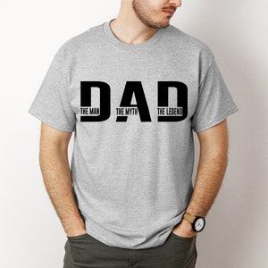 K&ouml;nnte beinhalten: Ein graumeliertes Kurzarm-T-Shirt mit dem schwarzen Schriftzug DAD in einer gro&szlig;en, fetten Schriftart. In den Buchstaben stehen die W&ouml;rter THE MAN, THE MYTH und THE LEGEND, wobei jeweils ein Ausdruck vertikal in jedem Buchstaben steht.