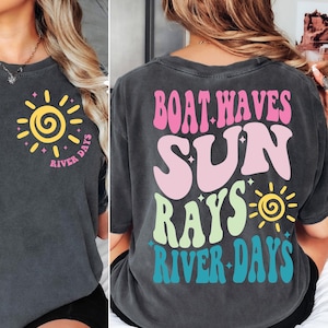 May include: A charcoal grey t-shirt featuring a small yellow sun graphic on the front chest with the text River Days. The back displays large, colourful, wavy text reading Boat Waves Sun Rays River Days with a yellow sun and small star accents.