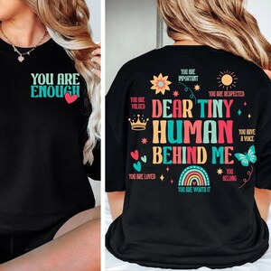 Könnte beinhalten: Schwarzes T-Shirt mit dem Text "You are enough" auf der Vorderseite und "Dear Tiny Human Behind Me" mit bunten Illustrationen auf der Rückseite.