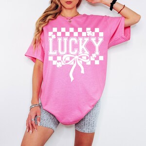 Pu&ograve; includere: Maglietta rosa oversize con un motivo a scacchi bianchi, un fiocco bianco e la parola "LUCKY" in lettere bianche.