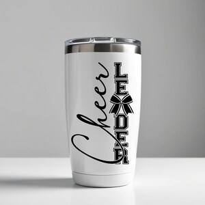 Puede incluir: Un vaso de acero inoxidable blanco con un dise&ntilde;o negro que dice "Cheer Leader" con un lazo negro en el medio de la palabra "Leader".
