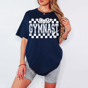 May include: A navy blue t-shirt with a white checkered pattern and the text "GYMNAST" in white with a white ribbon and a gymnast silhouette.