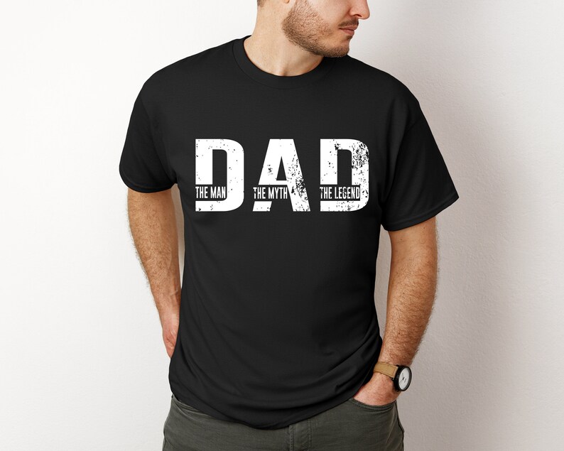 K&ouml;nnte beinhalten: Ein schwarzes Kurzarm-T-Shirt mit dem wei&szlig;en, im Used-Look gestalteten Schriftzug DAD in gro&szlig;en Druckbuchstaben. Unter jedem Buchstaben sind die W&ouml;rter THE MAN, THE MYTH und THE LEGEND in einer kleineren, klaren wei&szlig;en Schrift aufgedruckt.