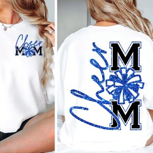 Peut inclure: Sweat-shirt blanc avec l'inscription "Cheer Mom" en lettres bleues pailletées. Le devant présente le texte "Cheer Mom" avec un motif de pompon bleu. Le dos du sweat-shirt porte le mot "Cheer" en cursive et les lettres "MOM" avec un motif de pompon bleu.