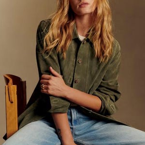 Può includere: Una persona indossa una giacca di camoscio verde, una camicia bianca e jeans azzurri, seduta su una sedia di legno. La giacca ha chiusure con bottoni e i jeans hanno un design a gamba larga. La persona indossa un anello.