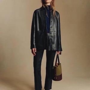 Könnte beinhalten: Schwarze Lederjacke mit Knopfleiste, getragen über einem marineblauen Rollkragenpullover und einer schwarzen Hose. Das Outfit wird durch schwarze Ankle Boots mit spitzer Zehenpartie und eine zweifarbige Handtasche in Olivgrün und Burgunderrot ergänzt.