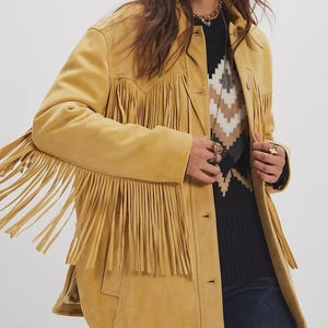 May include: Mustard yellow suede jacket with long fringe detailing on the sleeves, chest, and back. The jacket has a button-up front and a classic collar. The jacket is a statement piece, perfect for adding a touch of bohemian flair to any outfit.