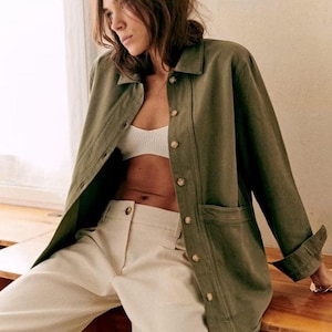 May include: An olive green jacket with large buttons and two front pockets. The jacket is paired with cream-colored pants and a white bra. The outfit is casual and stylish, suitable for everyday wear.
