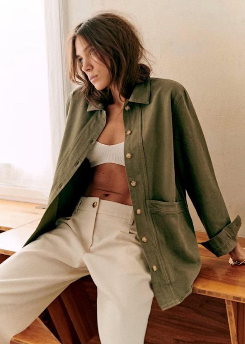 May include: An olive green jacket with large buttons and two front pockets. The jacket is paired with cream-colored pants and a white bra. The outfit is casual and stylish, suitable for everyday wear.