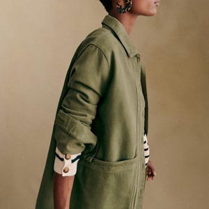 May include: An olive green jacket with a collared neckline and large front pockets. The jacket's sleeves are rolled up, revealing a striped sweater cuff. The model is wearing tortoise shell hoop earrings.