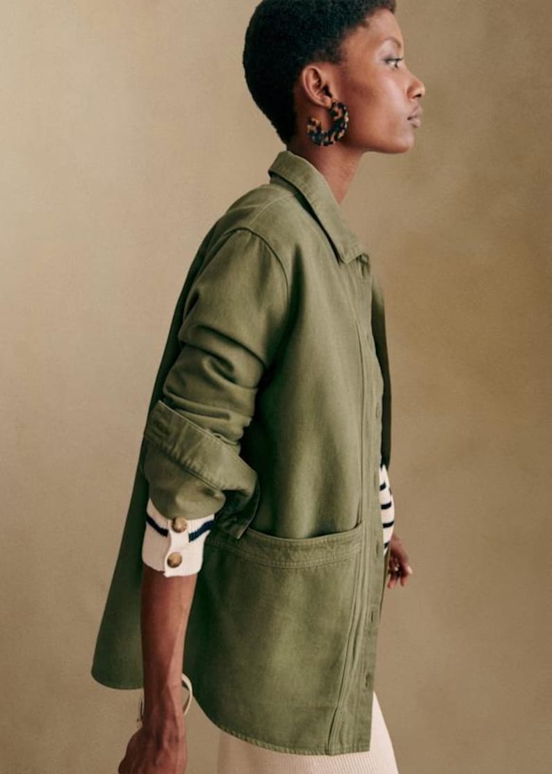May include: An olive green jacket with a collared neckline and large front pockets. The jacket's sleeves are rolled up, revealing a striped sweater cuff. The model is wearing tortoise shell hoop earrings.
