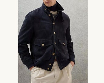 May include: A navy blue suede jacket with a stand-up collar and gold buttons. The jacket has two front pockets and is worn with cream-coloured trousers. The model is wearing a turtleneck sweater underneath.