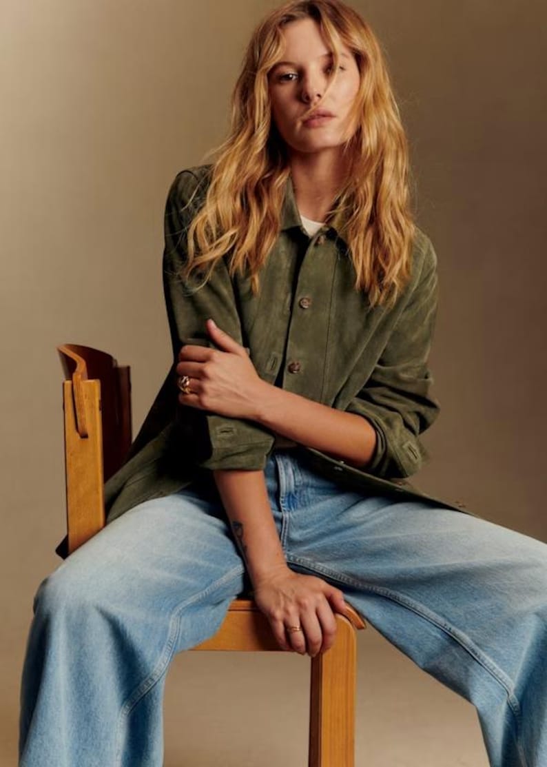 May include: A person wearing a green suede jacket, a white shirt, and light blue jeans, seated on a wooden chair. The jacket has button closures and the jeans have a wide leg design. The person is wearing a ring.