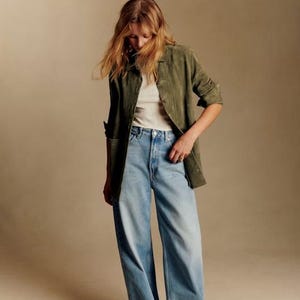 May include: A woman wearing a green suede jacket over a white ribbed tank top and light blue wide-leg jeans. The outfit is completed with brown and white sneakers. The background is a solid tan color.