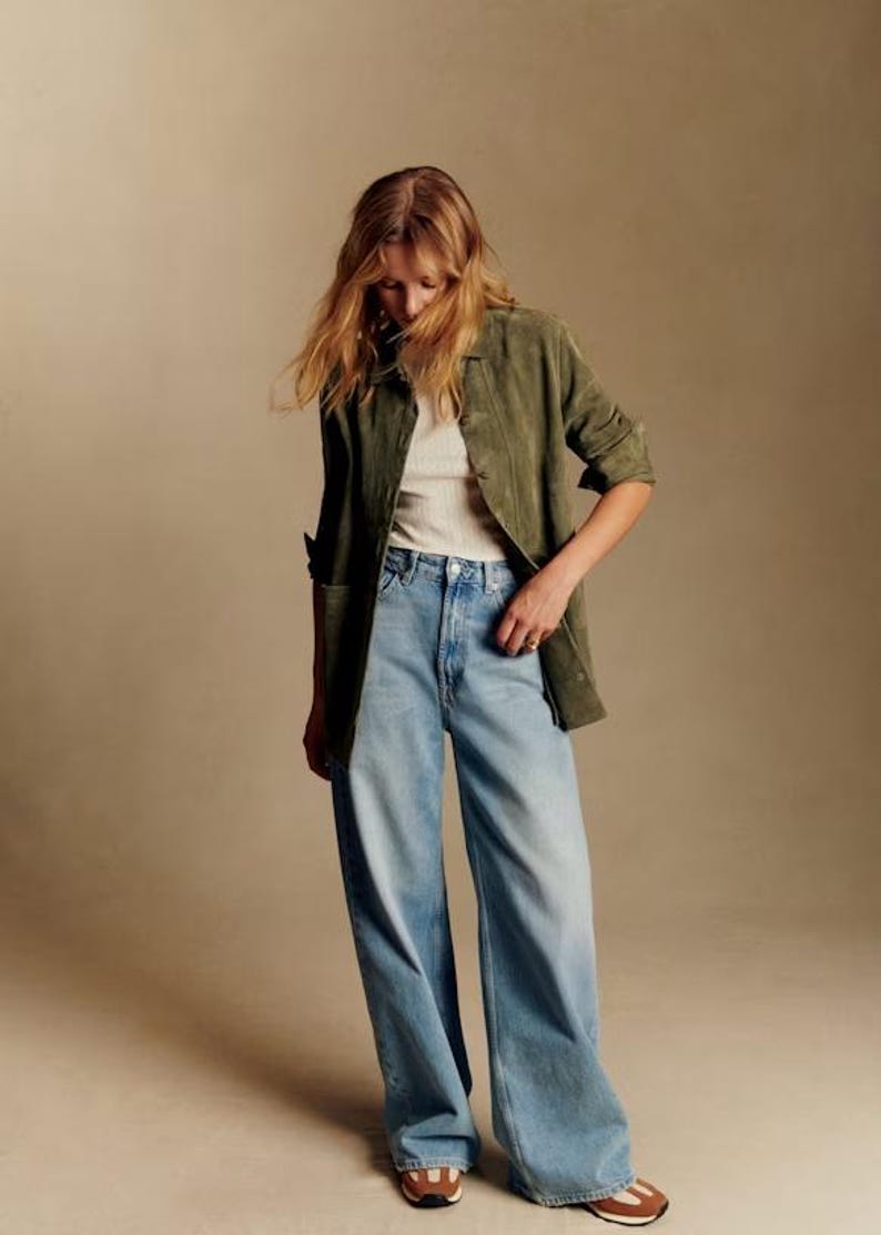 May include: A woman wearing a green suede jacket over a white ribbed tank top and light blue wide-leg jeans. The outfit is completed with brown and white sneakers. The background is a solid tan color.