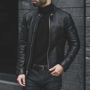 Könnte beinhalten: Schwarze Lederjacke mit Reißverschluss, getragen über einem schwarzen Rollkragenpullover. Die Jacke hat ein klassisches Design mit rundem Kragen. Die Person trägt auch einen schwarzen Gürtel und dunkelblaue Jeans.