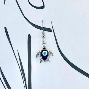 Puede incluir: Pendientes de plata con un pequeño dije de tortuga con un detalle de ojo azul malvado.
