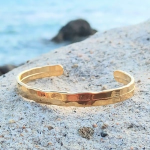 Puede incluir: Un brazalete de puño martillado de oro con una superficie texturizada. El brazalete está sobre una roca blanca con un fondo borroso de agua azul.