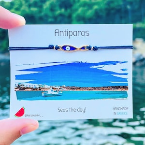 Peut inclure: Un bracelet en corde bleu marine avec un charm doré en forme d'œil bleu. Le bracelet est présenté sur une carte avec le mot "Antiparos" et une illustration de bord de mer. La carte contient également la phrase "Seas the day!"