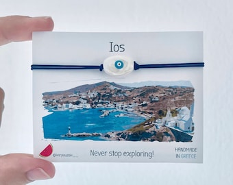 May include: A blue and white evil eye charm bracelet with a black elastic band. The bracelet is on a white card with the word "Ios" and the text "Never stop exploring! Handmade in Greece".