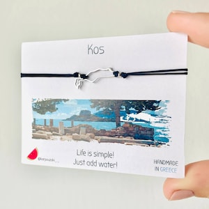 Può includere: Un braccialetto in corda nera con un ciondolo argentato a forma dell'isola di Kos. Il braccialetto è su una carta bianca con il testo "Kos" in alto, "Life is simple! Just add water!" al centro e "HANDMADE IN GREECE" in basso. Una piccola emoji di anguria si trova nell'angolo in basso a sinistra.