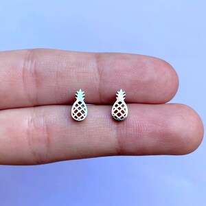 Puede incluir: Un par de pendientes de piña de plata. Los pendientes son pequeños y delicados, y son perfectos para añadir un toque de fantasía a cualquier atuendo.