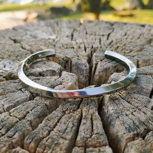 May include: A silver bangle bracelet with a slightly curved, flat design. The bracelet is resting on a rough, textured wooden surface.