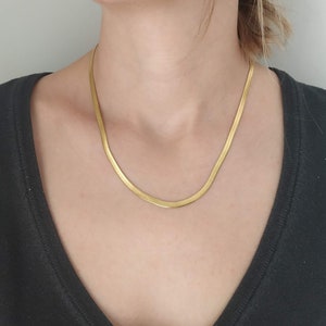 May include: A gold chain necklace with a flat, thin, and slightly curved design. The chain is worn by a person with a black shirt.