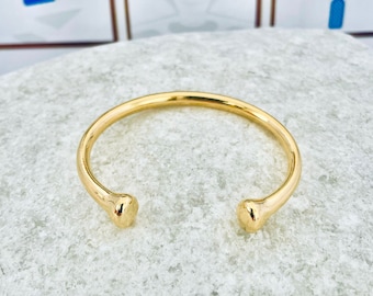 Könnte beinhalten: Ein goldenes Armreif-Armband mit abgerundeten Enden. Das Armband liegt auf einem weißen Stein.