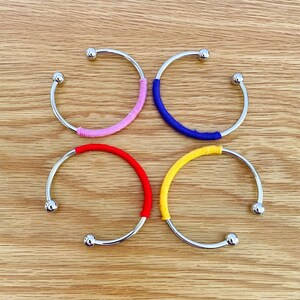 Könnte beinhalten: Vier silberne Armreifen mit farbigem Garn umwickelt. Die Armreifen sind quadratisch auf einer Holzoberfläche angeordnet. Die Farben des Garns sind rosa, blau, rot und gelb.