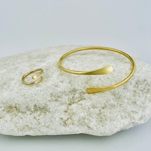 May include: A set of two gold-toned minimalist jewelry pieces: a thin bangle bracelet with a curved, open design and a simple ring with a similar curved shape.