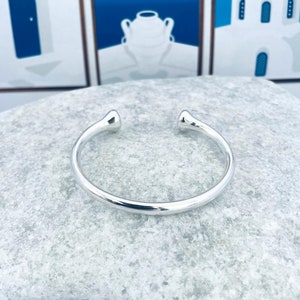 May include: A shiny silver cuff bracelet with rounded ends. The open design allows for easy wear. The bracelet is displayed on a light gray, textured surface, with a blurred background featuring blue and white artwork.