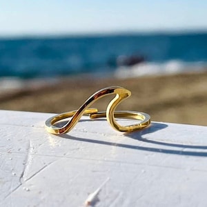 Op de afbeelding: Een gouden golfrijng, een simpel en delicaat sieraad, wordt getoond op een wit oppervlak met een wazige achtergrond van een strand en oceaan.
