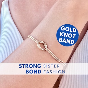 Puede incluir: Un brazalete dorado con un nudo se muestra en un brazo. El brazalete presenta un diseño de nudo en el centro. El texto "GOLD KNOT BAND" está en un círculo azul. El texto "STRONG SISTER BOND FASHION" está debajo.