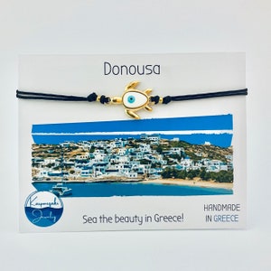 May include: A gold-coloured turtle charm bracelet with a blue eye design, strung on black cord. The bracelet is displayed on a card with the word "Donousa" and a scenic image of a Greek island. The card also includes the text "Sea the beauty in Greece!"