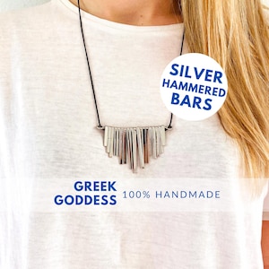 Op de afbeelding: Een zilveren ketting met gehamerde staafhangers hangt aan een wit shirt. De ketting is gelabeld met "Silver Hammered Bars" en "Greek Goddess 100% Handmade".