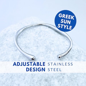 Può includere: Un bracciale a fascia color argento con una finitura liscia e lucida e le estremità arrotondate. Il testo GREEK SUN STYLE è stampato in un cerchio blu, e il testo ADJUSTABLE STAINLESS STEEL è stampato in blu nella parte inferiore.