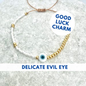 May include: White and gold beaded bracelet with a blue evil eye charm. The bracelet is tied with a brown cord and has the text "GOOD LUCK CHARM" and "DELICATE EVIL EYE" on the image.